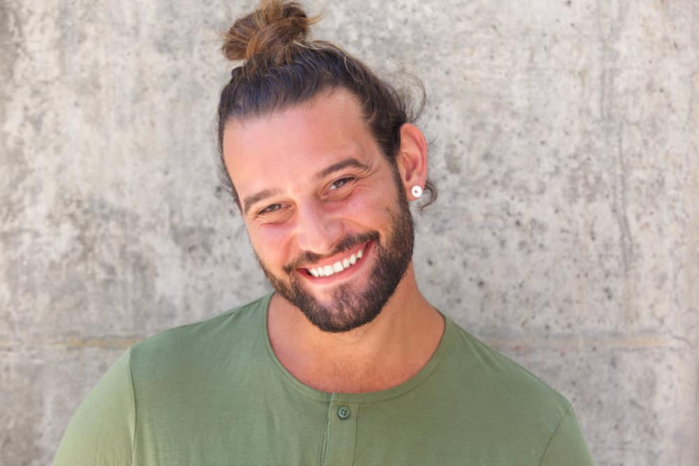 A white with facial hair wearing a messy man bun hairstyle undercut with a half-up top knot (aka bro bun) on his brown and blonde thin hair.