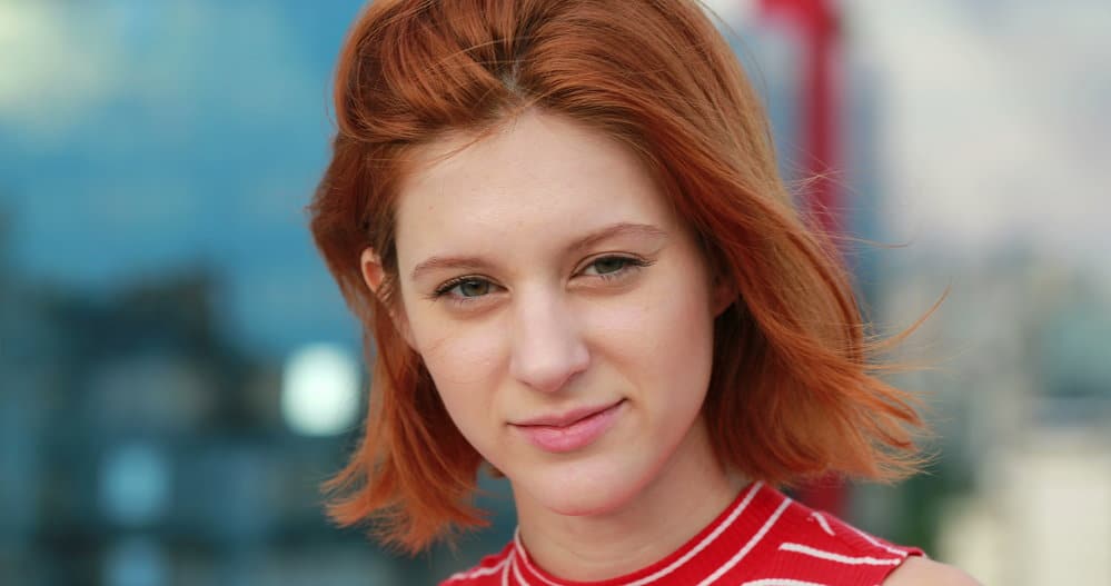 A super stylish young woman with red straight hair wearing a short hairstyle with blunt bangs styled with an organic holding spray on her oval face shape.