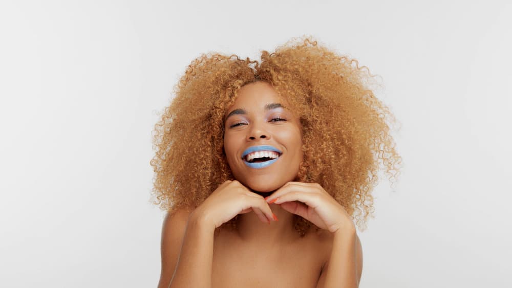 A beautiful black female rocks her curly hair colored with medium brown hair dye, which is a few shades darker than her natural hair color, and adds a pop of color with blue lipstick and orange nail polish.