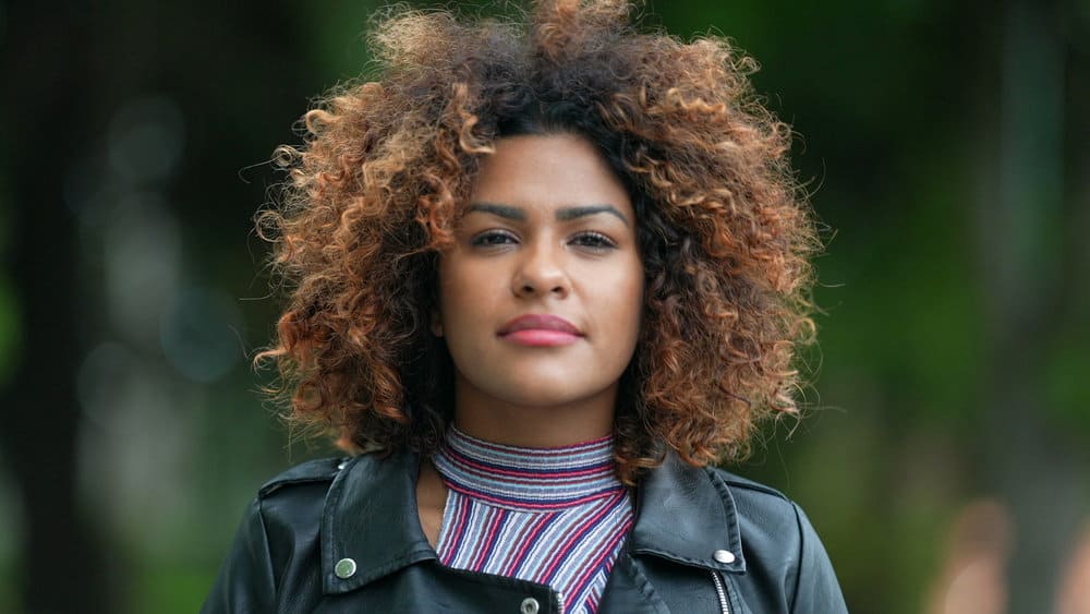 A stunning black female with reddish-brown skin showcases her perfect skin and face shape, highlighted by her dark brown hair mixed with rose gold dyed hair strands.