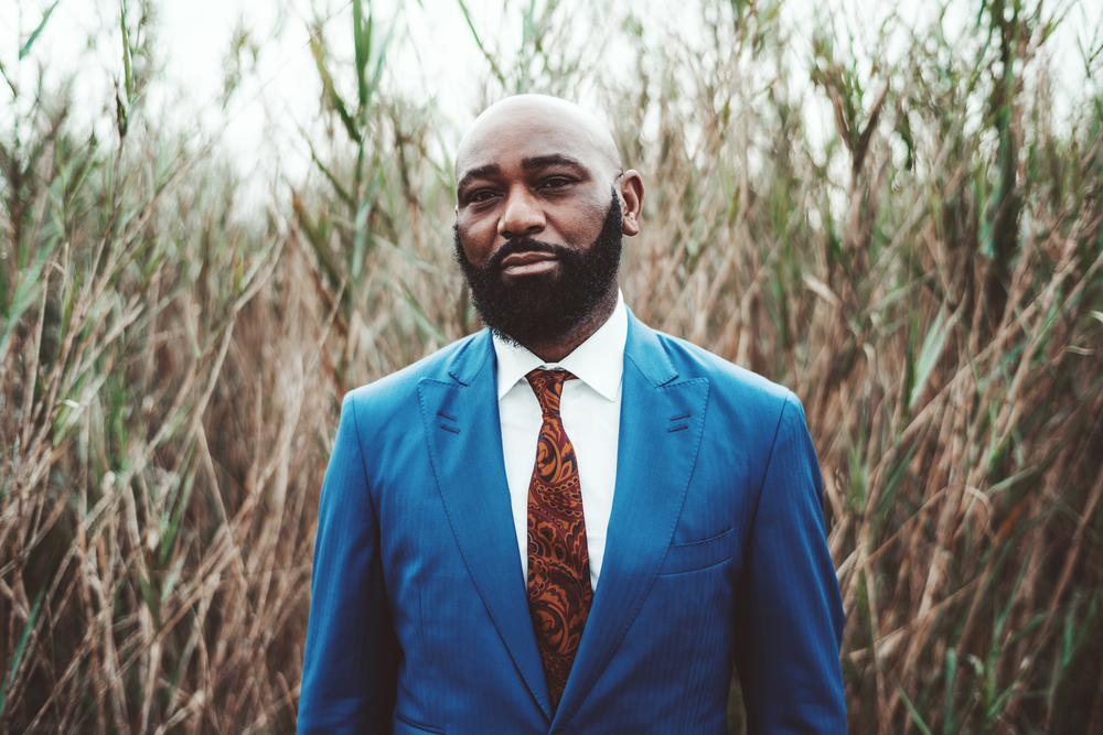 A handsome African man with a shaved head due to balding hair on his head and thick, dark brown facial hair creating a very masculine hairstyle.