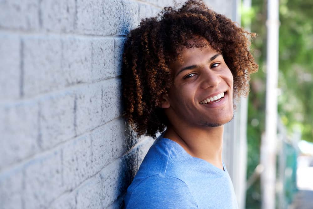 A young black man with fine hair is wearing his curly long hair in a classic style that resembles a dark brown and tan ombre hairdo with natural curls.
