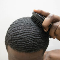 A young African American man with short hair and a 180 wave pattern on his type 4 hair texture is using a boar bristle brush on his wide head shape.