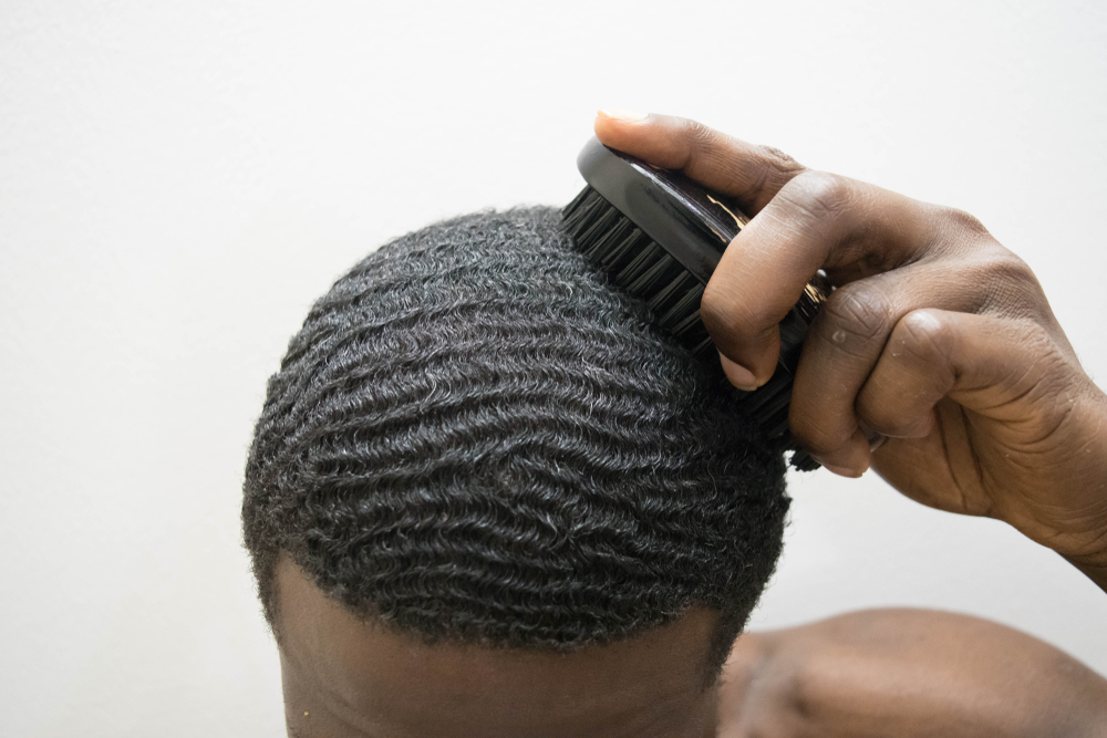A young African American man with short hair and a 180 wave pattern on his type 4 hair texture is using a boar bristle brush on his wide head shape.
