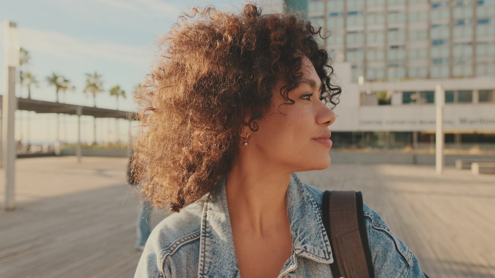 A black woman with a cute, confident smile proudly shows off her natural curls styled with shea butter and natural oils, like coconut oil, to hydrate her frizzy hair strands.