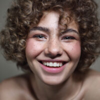 A beautiful young freckled girl with long curly hair and shaggy bangs in a stunning curly pixie cut style that perfectly highlights her oval face shape.