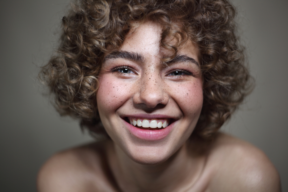 A beautiful young freckled girl with long curly hair and shaggy bangs in a stunning curly pixie cut style that perfectly highlights her oval face shape.