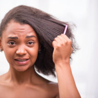 A disappointed black female with matted hair strands is combing through her hair with a wide-toothed comb after using detangling spray on lightly dampened hair.