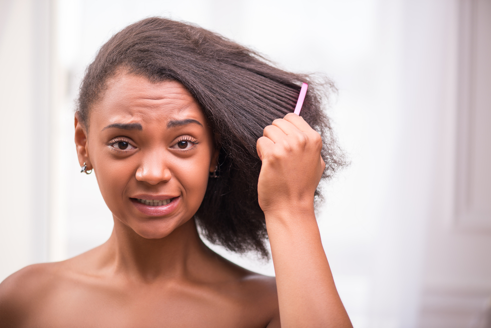 A disappointed black female with matted hair strands is combing through her hair with a wide-toothed comb after using detangling spray on lightly dampened hair.