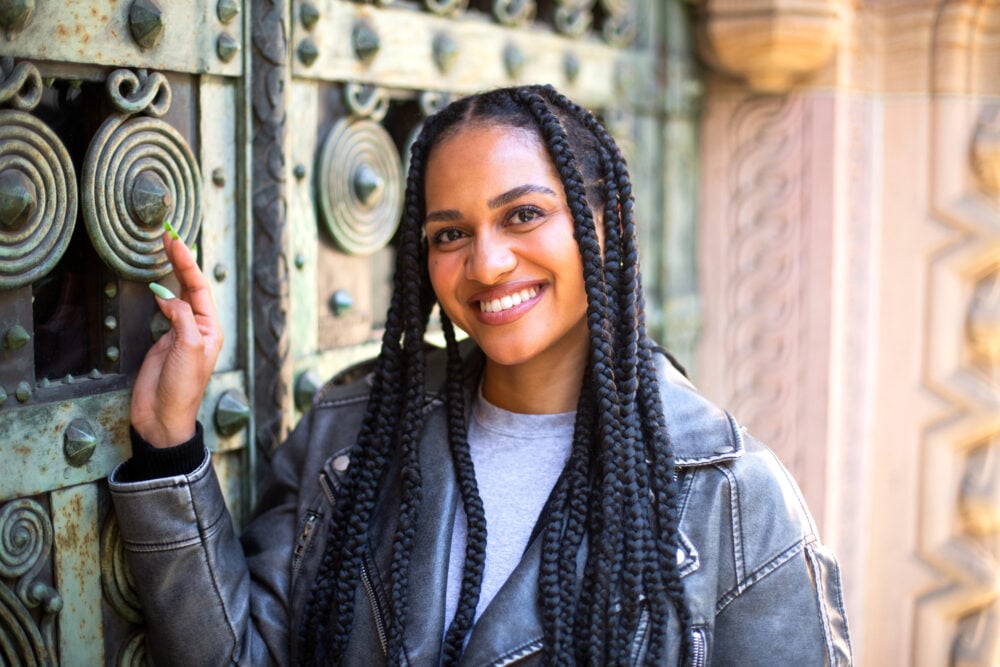 Smiling woman with large knotless braids and a center part, resting one hand on an ornate bronze door
