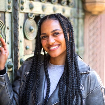 Smiling woman with large knotless braids and a center part, resting one hand on an ornate bronze door
