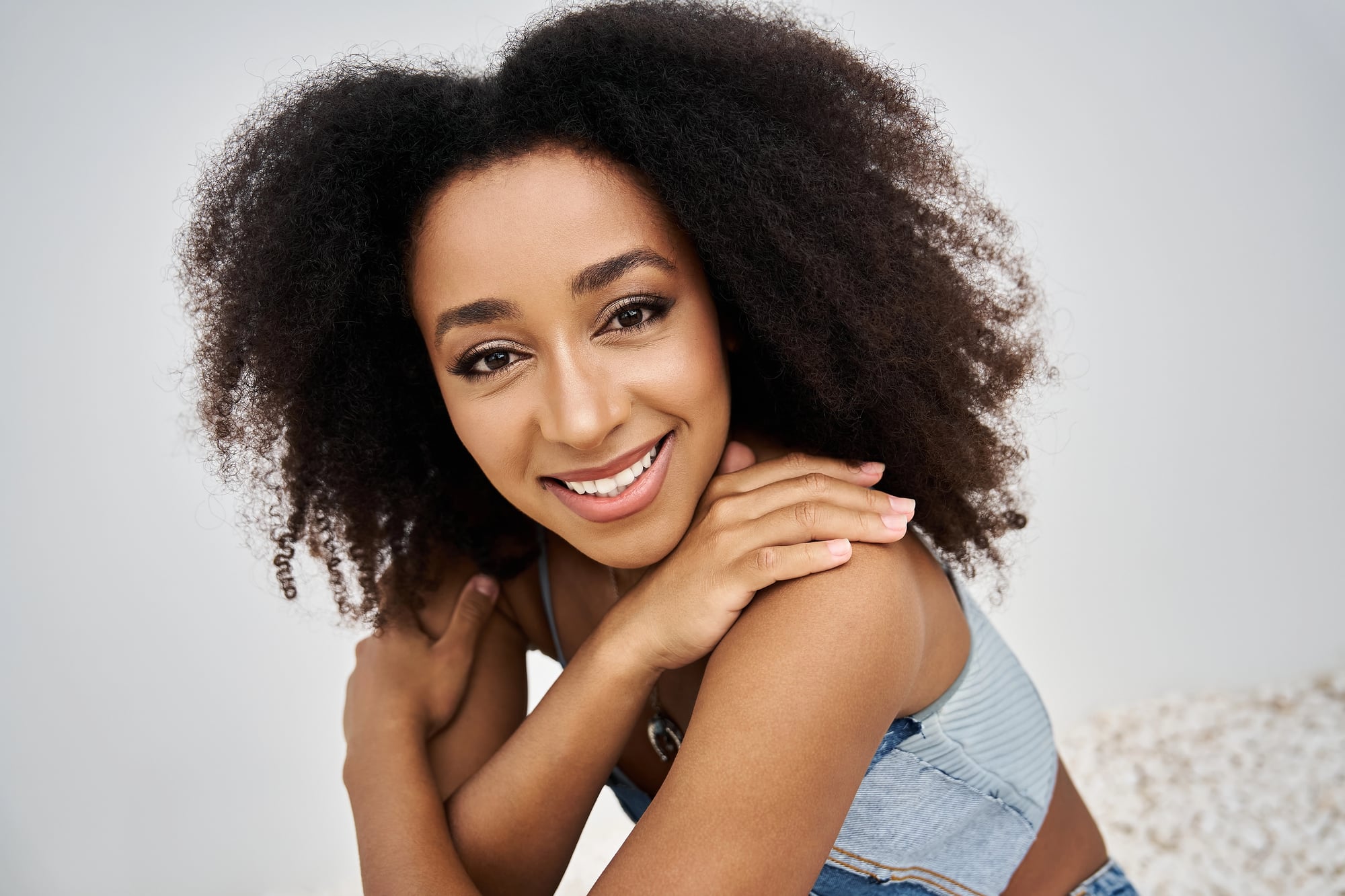 A young Black woman with a full type 4 afro resting her hands on her shoulder and smiling