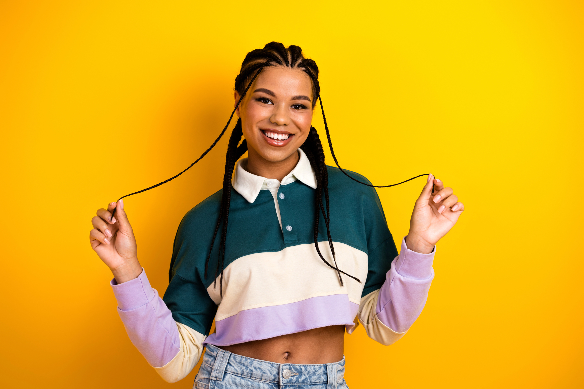A young Black woman with long cornrow braids holding two of them out to either side, smiling, against a bright yellow background