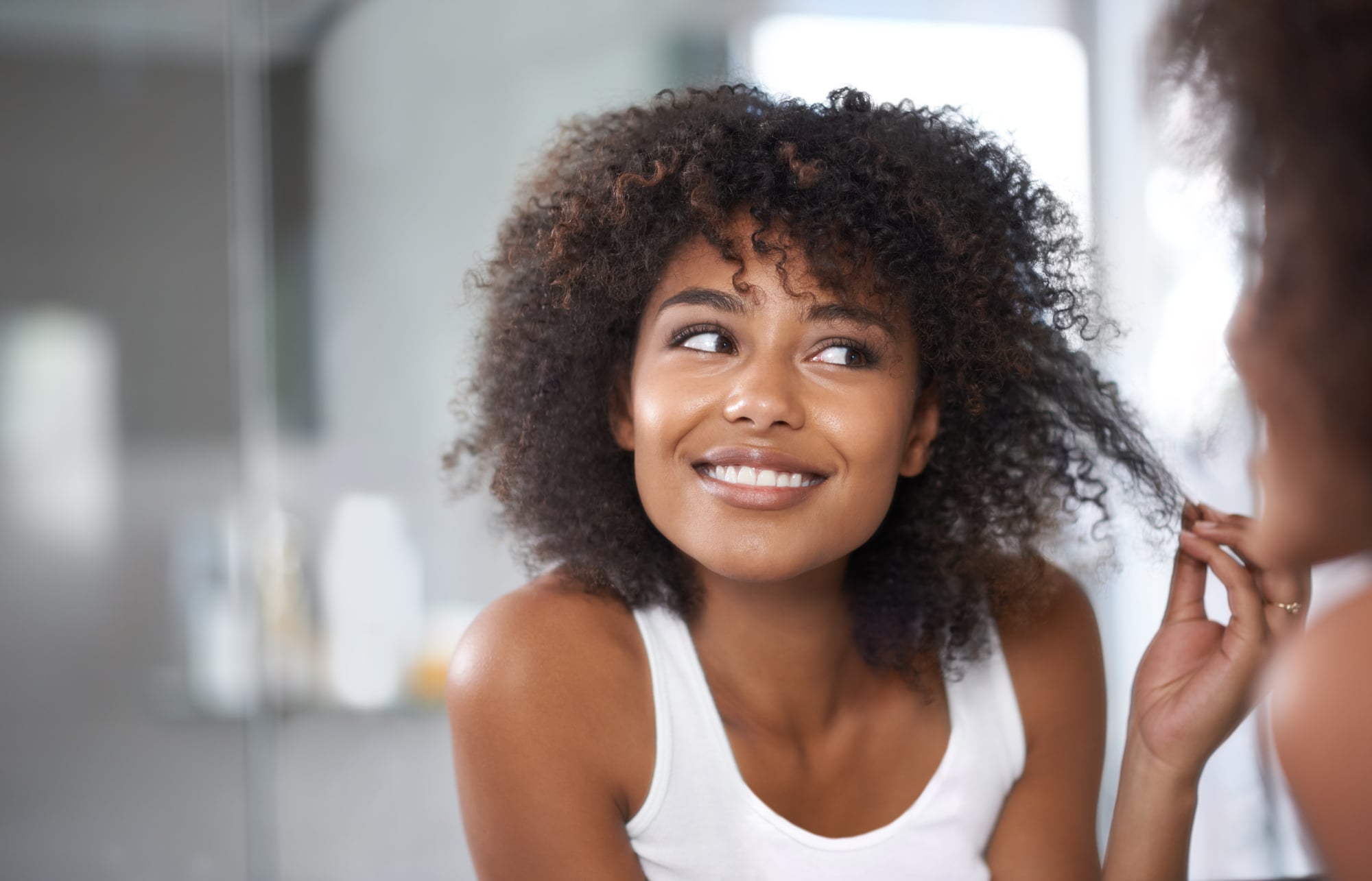 Shedding vs Breakage: How to Tell the Difference 2 The same woman holding one section of her curls out from her head at the bathroom mirror, inspecting the strand