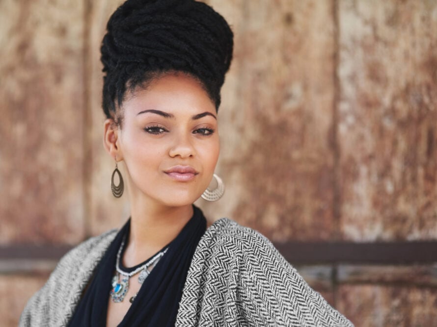 Woman with long box braids gathered into a high bun, standing in front of a weathered wooden wall