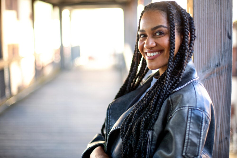 Woman on a covered wooden footbridge, her knotless braids lying flat at the roots and falling over her jacket