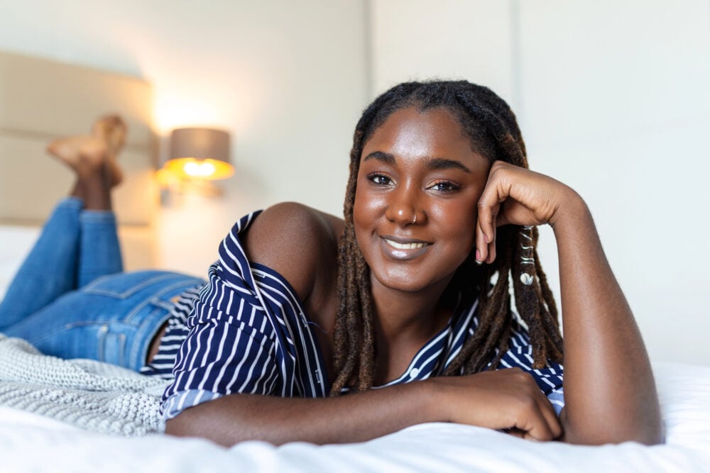A woman lying on her front across a white bed, chin propped on one hand, locs falling over her shoulder