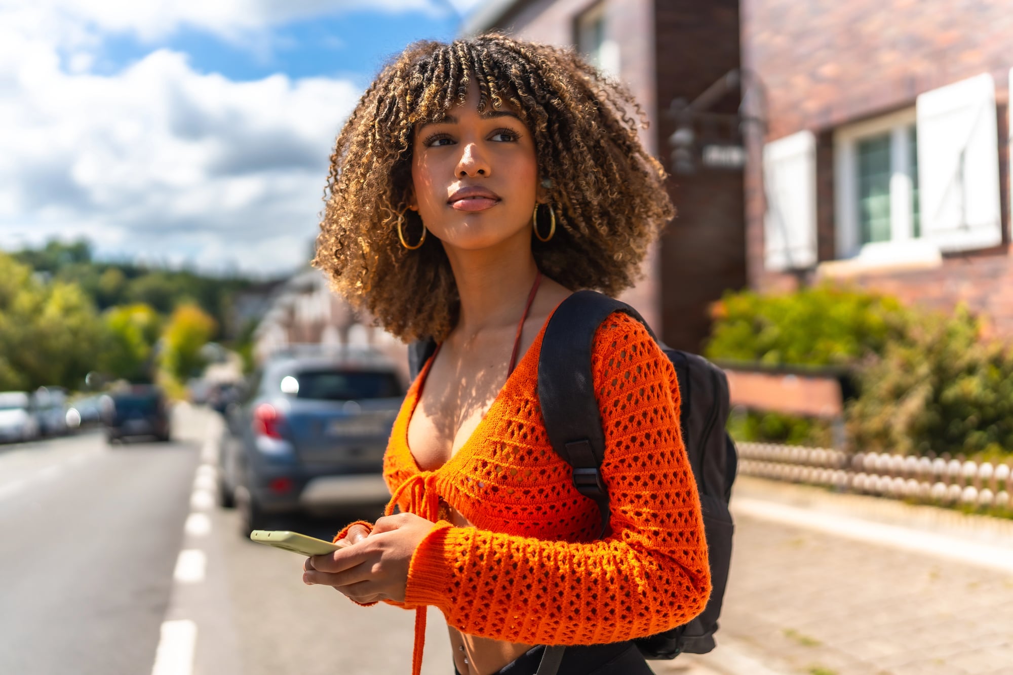 A young woman with a curly bob and blonde highlights standing on a sunlit street, holding a phone