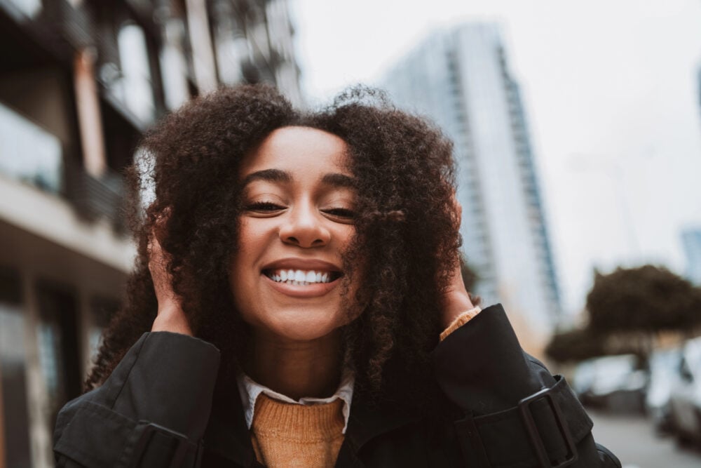 Black woman laughing with both hands lifted into her shoulder-length curls on a city street