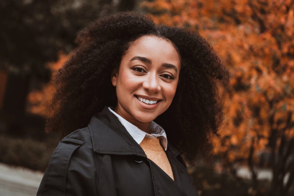 Black woman with a full, defined curly afro smiling outdoors in a black trench coat, with blurred orange autumn leaves behind her
