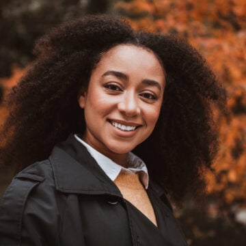 Black woman with a full, defined curly afro smiling outdoors in a black trench coat, with blurred orange autumn leaves behind her
