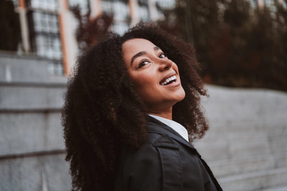 Black woman tilting her head back and laughing, her long curls falling loose over a black coat