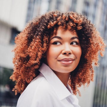 A woman with a rounded copper-brown curly afro and darker roots, in a white shirt, looking into the camera