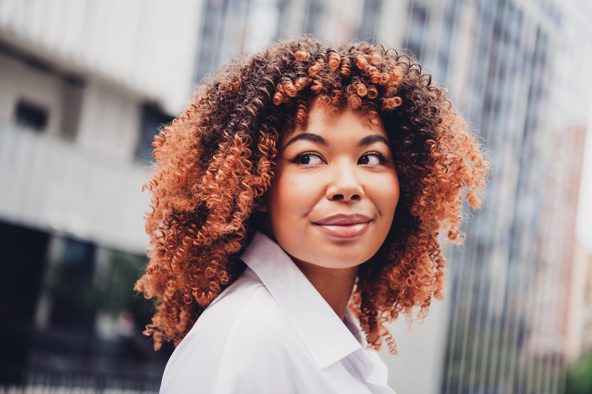 A woman with a rounded copper-brown curly afro and darker roots, in a white shirt, looking into the camera