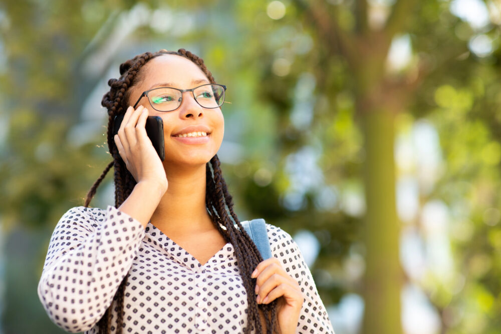 Woman with long box braids and glasses smiling while talking on her phone outdoors