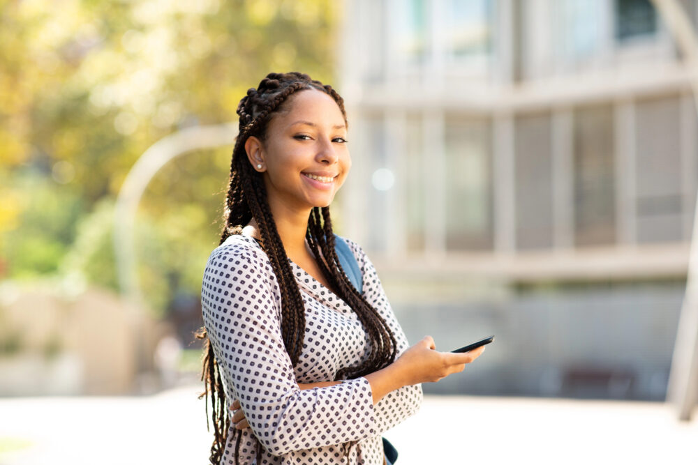 Young Black woman with long box braids past her waist, smiling outdoors while holding her phone