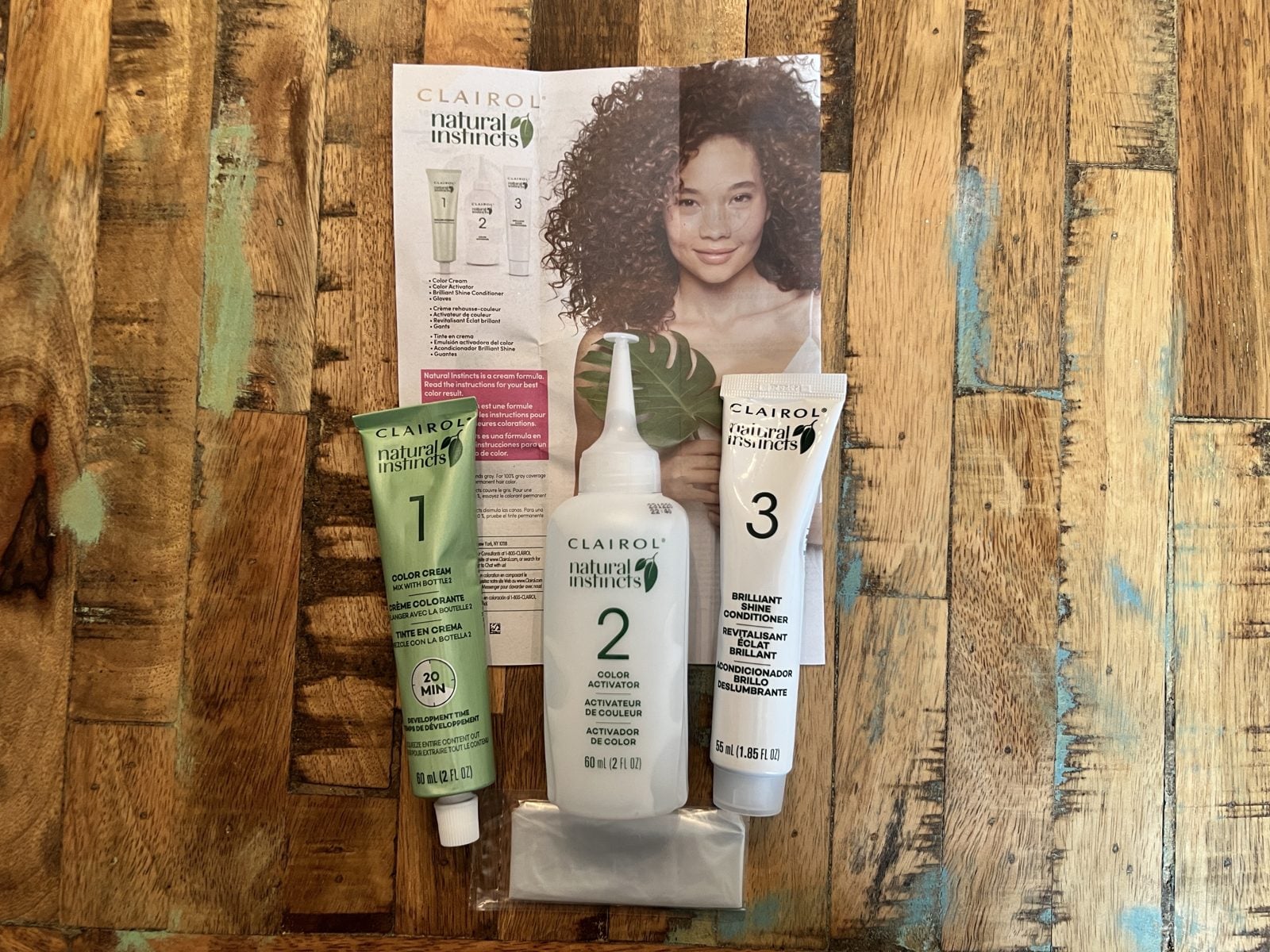 Clairol Natural Instincts kit laid out on a wood table: box, color creme, activator bottle, and conditioner tube
