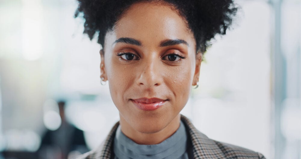 Black woman with a short natural afro smiling in a houndstooth blazer, with a softly blurred office behind her