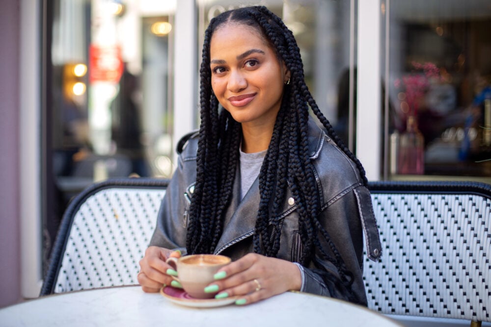 Woman at an outdoor café table holding a cup of coffee, her long knotless braids falling past her shoulders