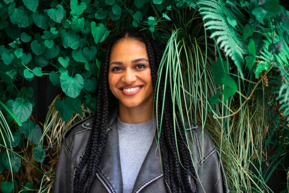 Woman smiling in front of a wall of ivy and ferns, with her knotless braids parted down the center