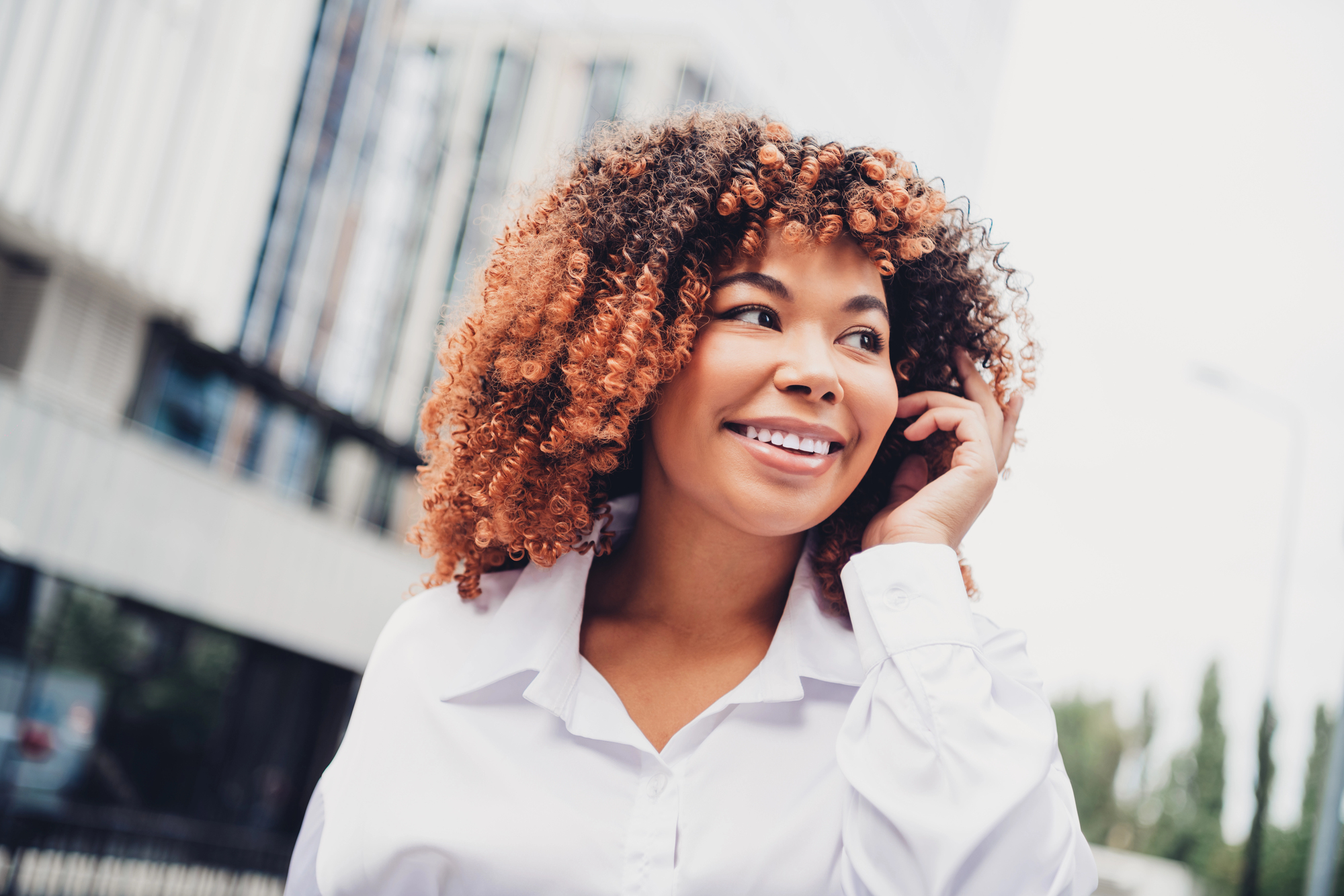 The same woman laughing with one hand lifted to her copper curls, glass office buildings behind her