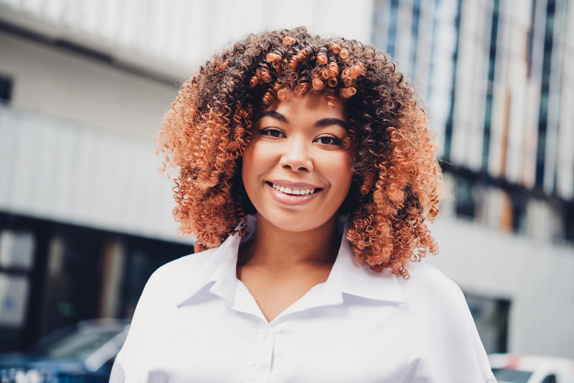 The same woman smiling on a city street in a white shirt, her copper-brown curls lit from behind