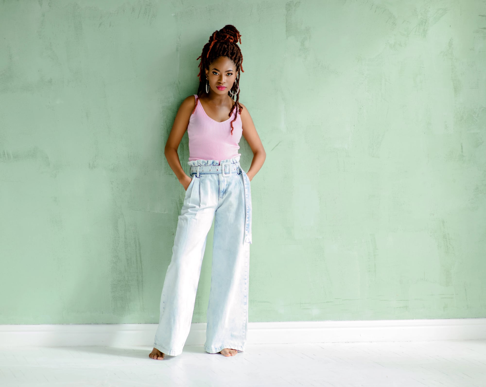 A woman standing barefoot, hands in her pockets, auburn locs in a high bun, in a pink tank and wide-leg jeans