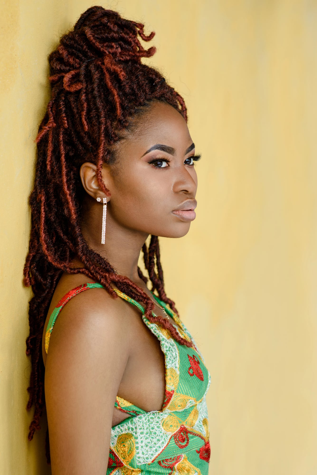 A woman in profile against a yellow wall, long auburn locs pulled half up over a braided base, in a green print dress