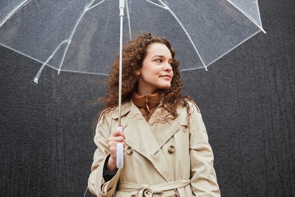The same woman with curly hair standing under a clear umbrella in front of a dark wall