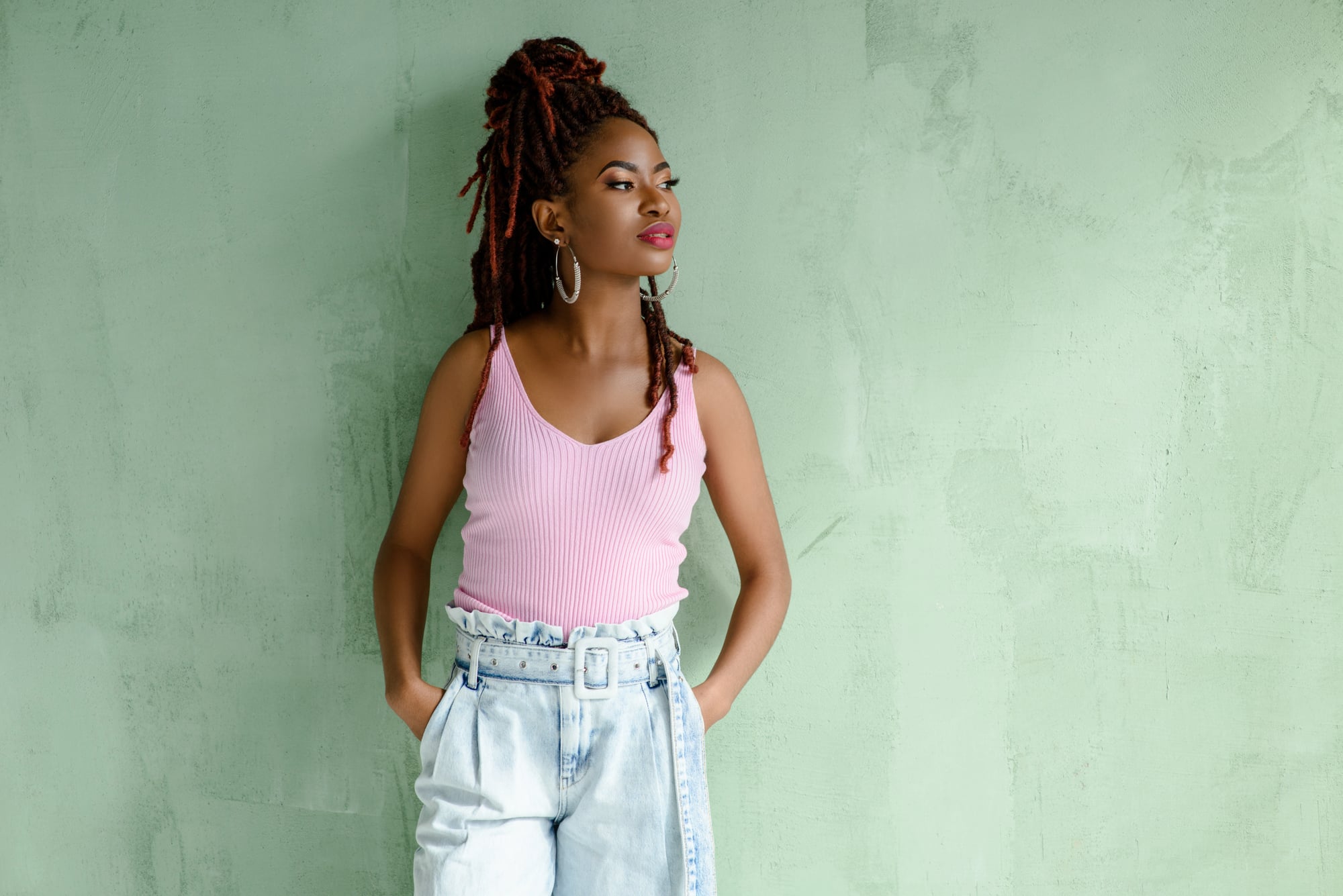 A woman leaning on a green wall and looking to one side, auburn locs gathered high with two falling loose at her face