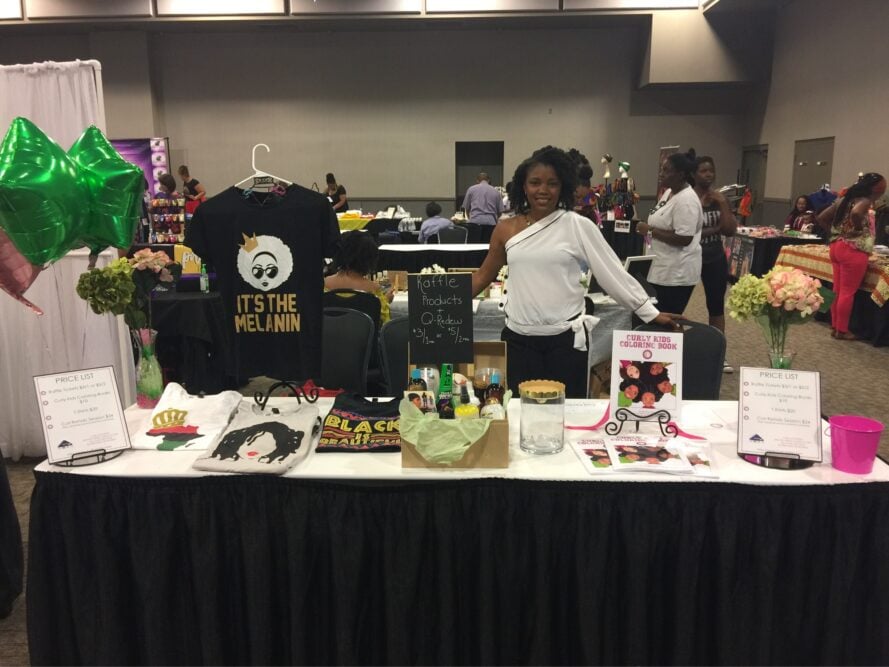 Kira Byrd standing behind her hair show booth table with copies of Curly Kids Coloring Book, T-shirts, and hair products