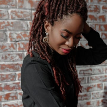 A woman looking down with one hand behind her head, auburn locs parted into neat rows at the scalp, against a brick wall