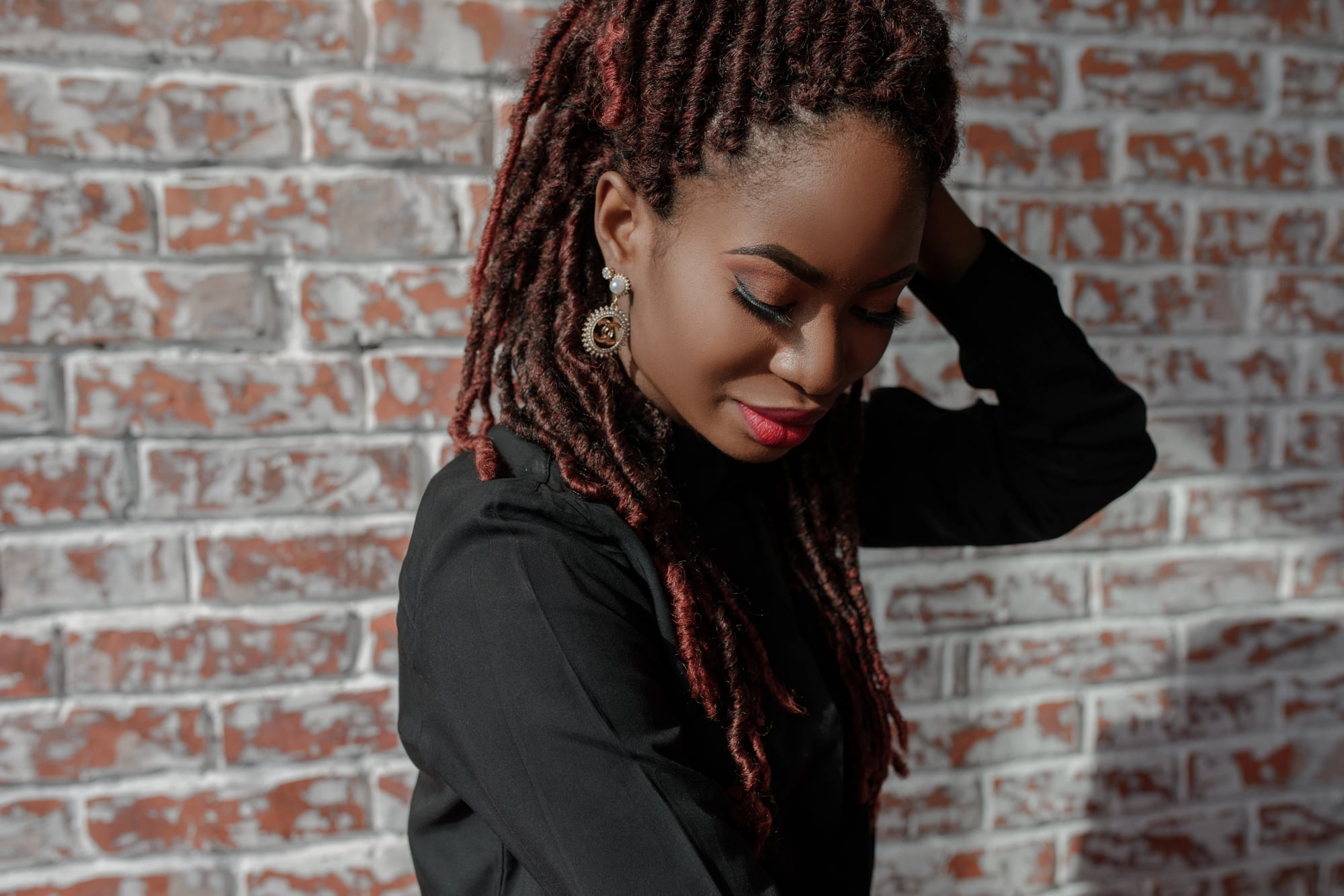 A woman looking down with one hand behind her head, auburn locs parted into neat rows at the scalp, against a brick wall