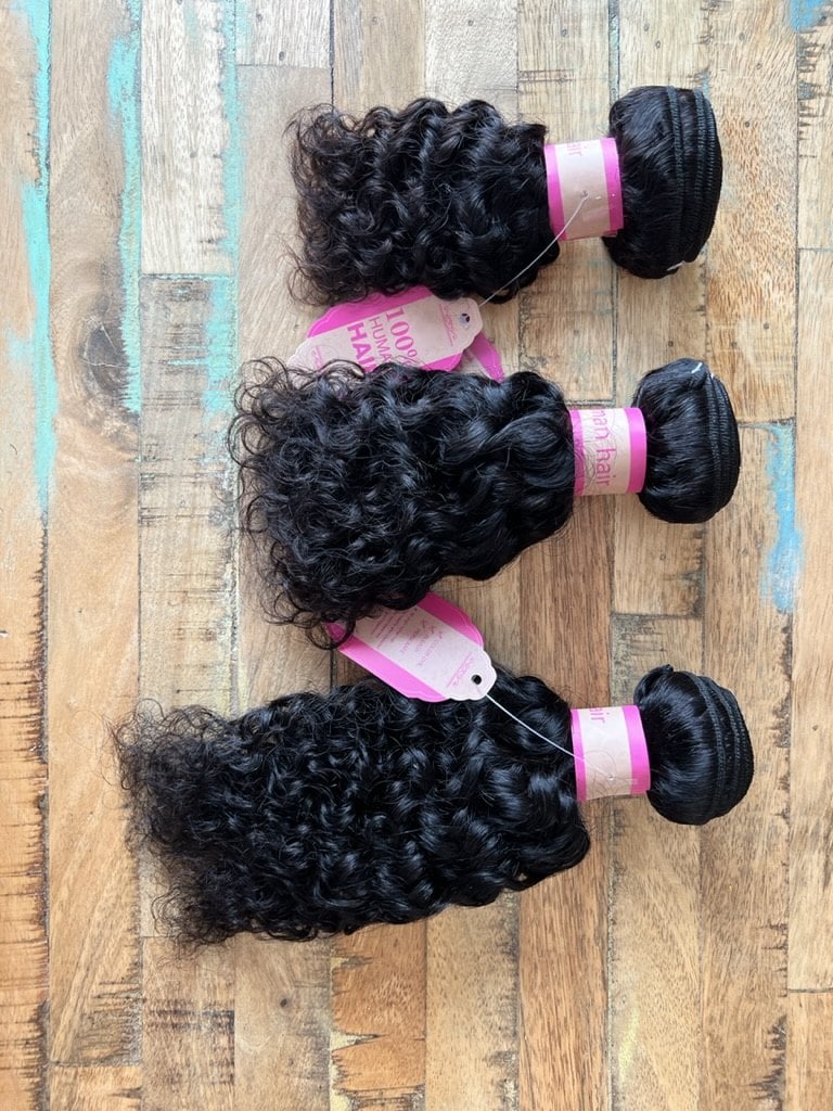 Three bundles of black curly weave hair with pink bands and tags, lined up on a wood table